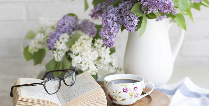 En uppfälld bok, ett par glasögon och en gammaldagskaffe kopp står på en bricka. På bordet står även en vas med lila syrener. 