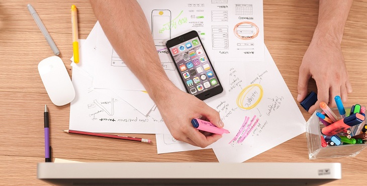 Två händer som håller i en rosa markeringspenna. En telefon och papper på ett bord syns och ska illustrera att ett projekt pågår. 