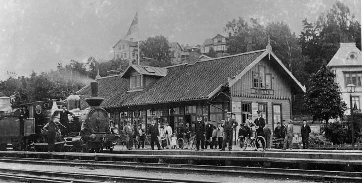 Många människor står utanför ett gammalt stationshus i trä.